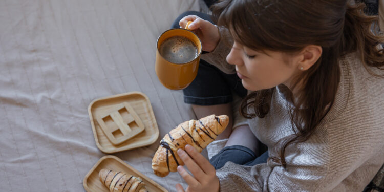 Young woman wrapped in a plaid drinking coffee with croissant in bed, top view, concept of delicious breakfast at home.